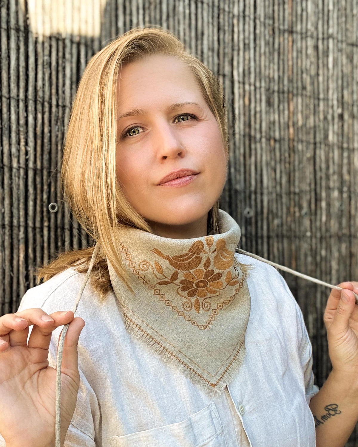 A woman wearing a beige linen neck gaiter with brown floral embroidery, holding the ties against a rustic bamboo fence background.