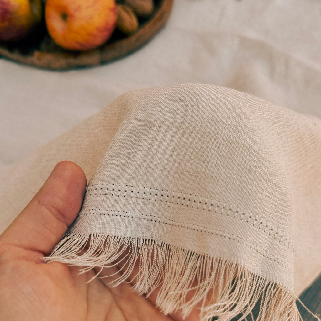 A close-up of a hand holding a light beige woven textile with fringe and embroidered dotted trim, with a softly blurred background showing fabric and fruit.