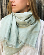 A woman wearing a pale sage green linen scarf with fringed edges, styled over a light-colored linen shirt against a rustic wooden background.