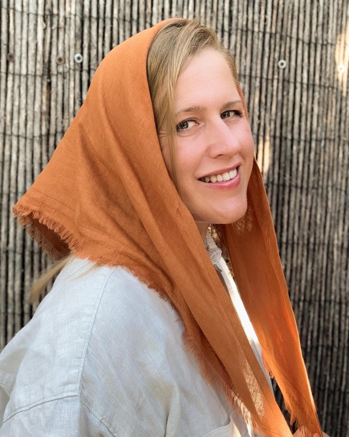 A smiling woman wearing a rust-orange linen headscarf with frayed edges over a light-colored shirt, standing in front of a dark woven bamboo screen.