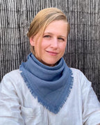 A woman with blonde hair tied back, wearing a dusty blue fringed triangle scarf over a white linen shirt, standing in front of a rustic bamboo fence.