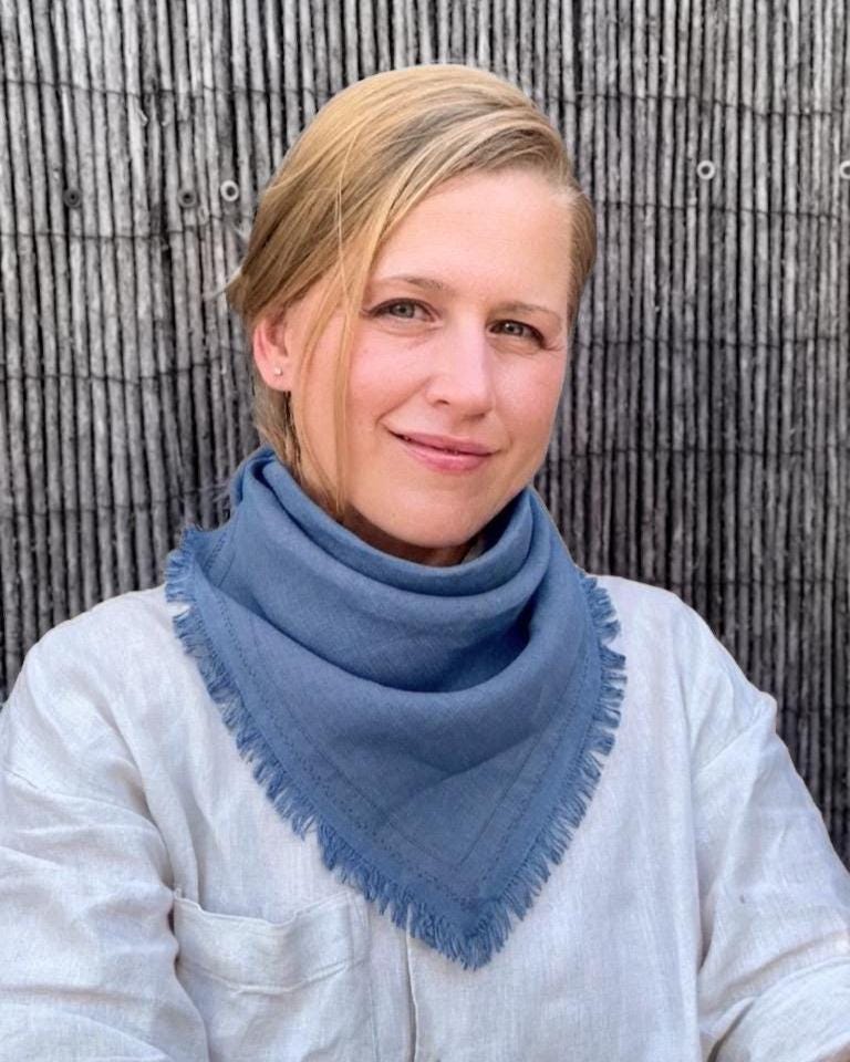 A woman with blonde hair tied back, wearing a dusty blue fringed triangle scarf over a white linen shirt, standing in front of a rustic bamboo fence.