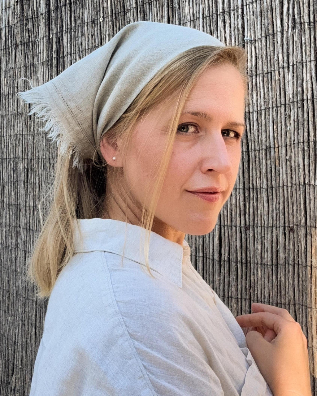 A woman wearing a light beige linen headscarf and matching linen shirt, posing in front of a rustic wooden fence.