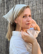 A woman wearing a light beige linen headscarf and a white linen shirt, posing in front of a vertical wicker fence.