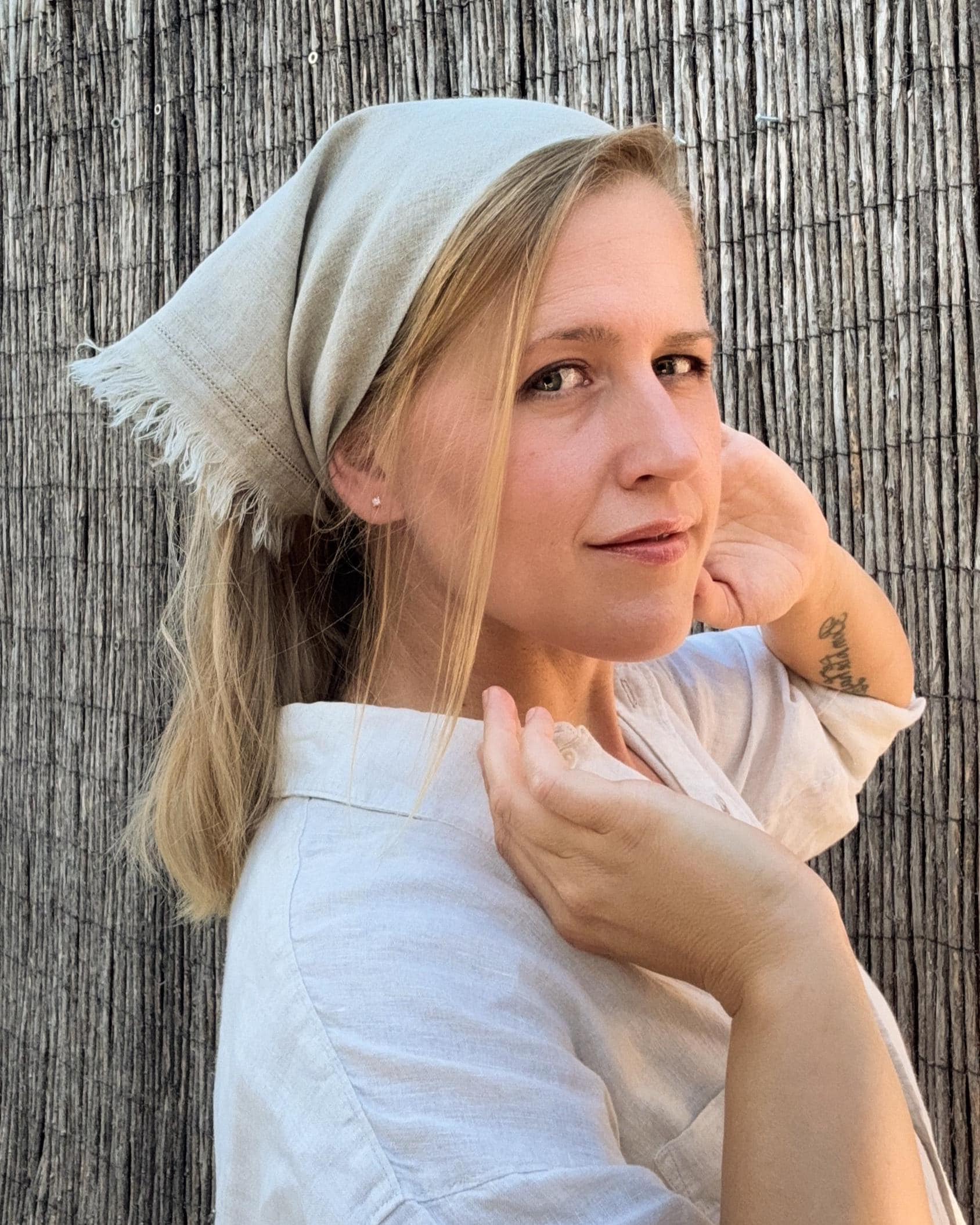 A woman wearing a light beige linen headscarf and a white linen shirt, posing in front of a vertical wicker fence.
