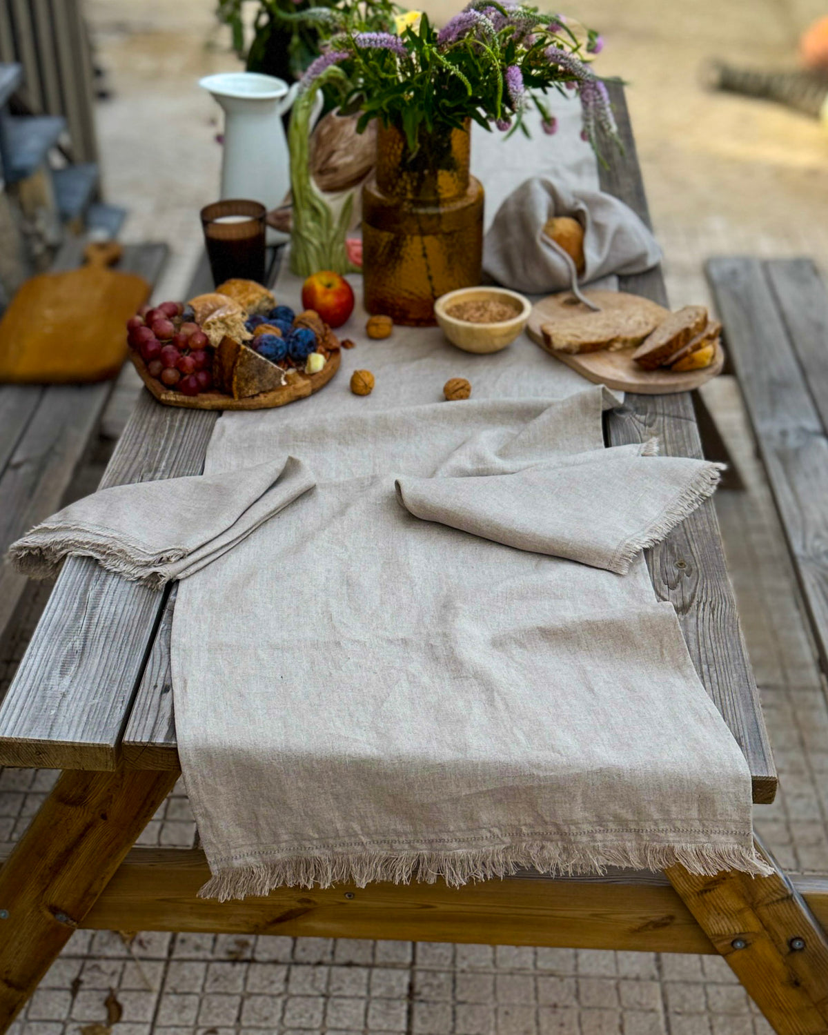 A light beige linen tablecloth with fringed edges draped over a rustic wooden picnic table, styled with bread, grapes, flowers, and a small bowl in an outdoor setting.