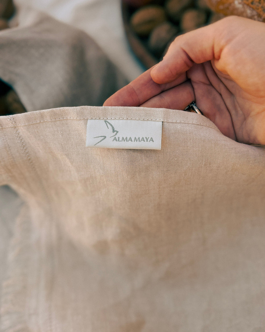 A close-up of a hand holding a light beige linen garment, showing a white fabric label with the brand name 'ALMA MAYA' printed on it.