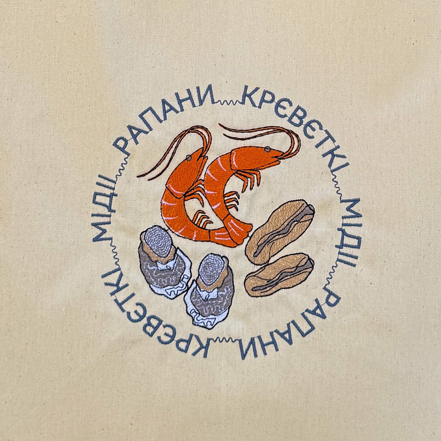 A beige canvas tote bag with two shoulder straps, featuring a circular graphic embroidered in the center with a cartoon shrimp, blue and brown shoes, and blue Cyrillic text, displayed against a brown woven straw background.