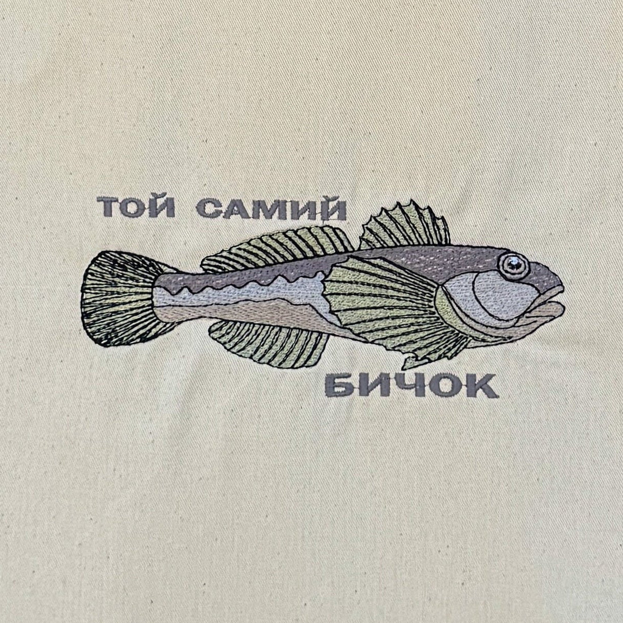 A natural canvas tote bag with two shoulder straps, featuring a embroidered illustration of a fish and the text 'той самий' and 'бичок' on the front, hanging against a dark brown bamboo reed wall.