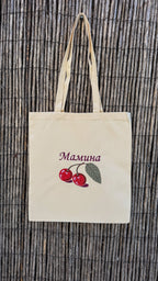 A natural canvas tote bag with two handles, featuring an embroidered design of two red cherries with green leaves and the Ukrainian word 'Мамина' in purple. The bag is displayed flat against a brown bamboo reed background.