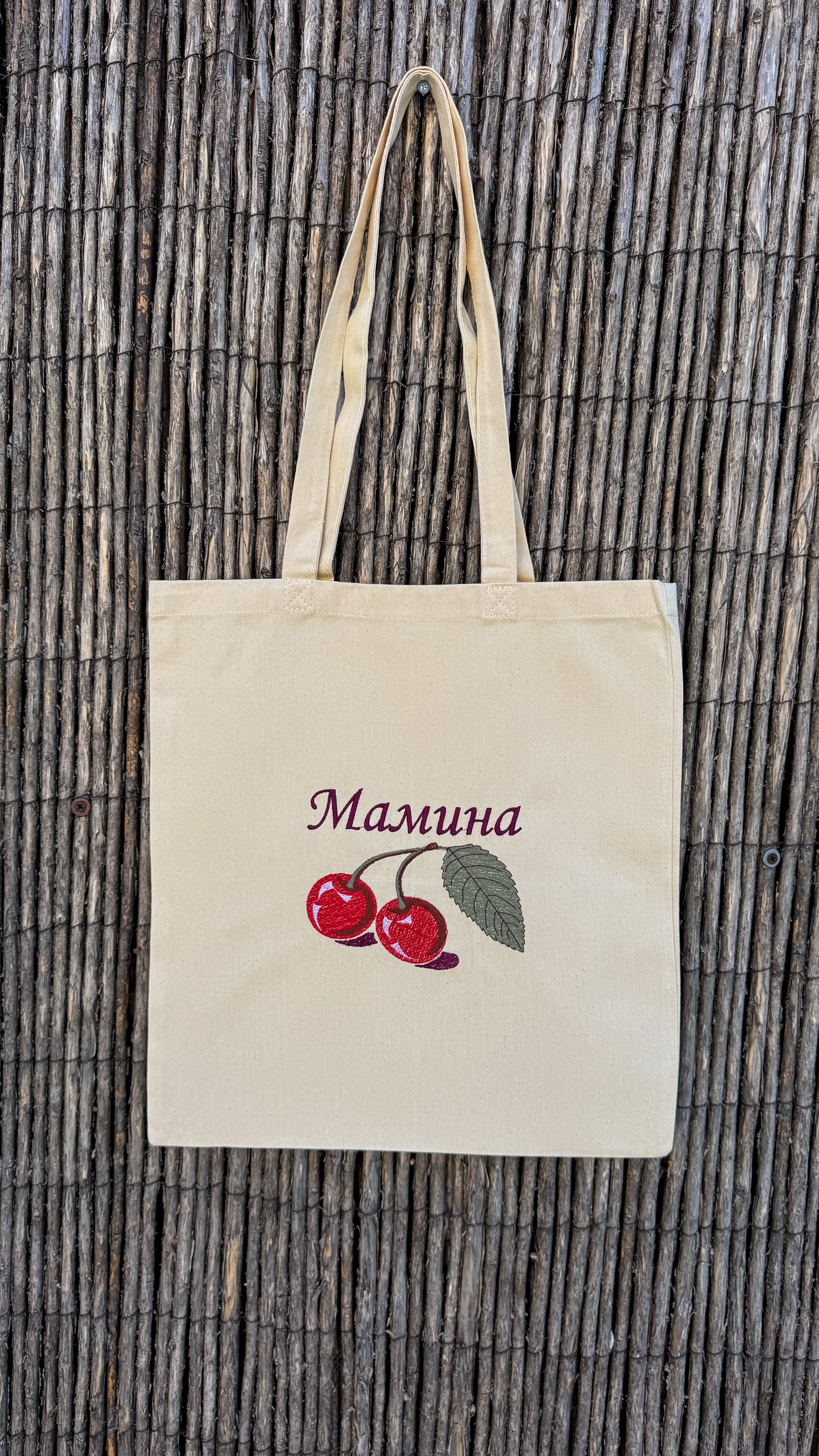 A natural canvas tote bag with two handles, featuring an embroidered design of two red cherries with green leaves and the Ukrainian word 'Мамина' in purple. The bag is displayed flat against a brown bamboo reed background.