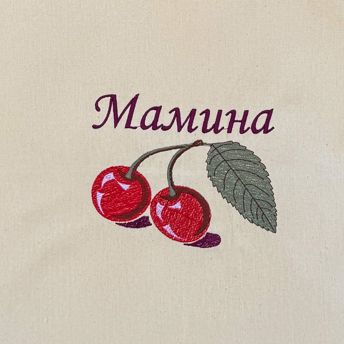 A natural canvas tote bag with two handles, featuring an embroidered design of two red cherries with green leaves and the ukrainian word 'Мамина' in purple. The bag is displayed flat against a brown bamboo reed background.