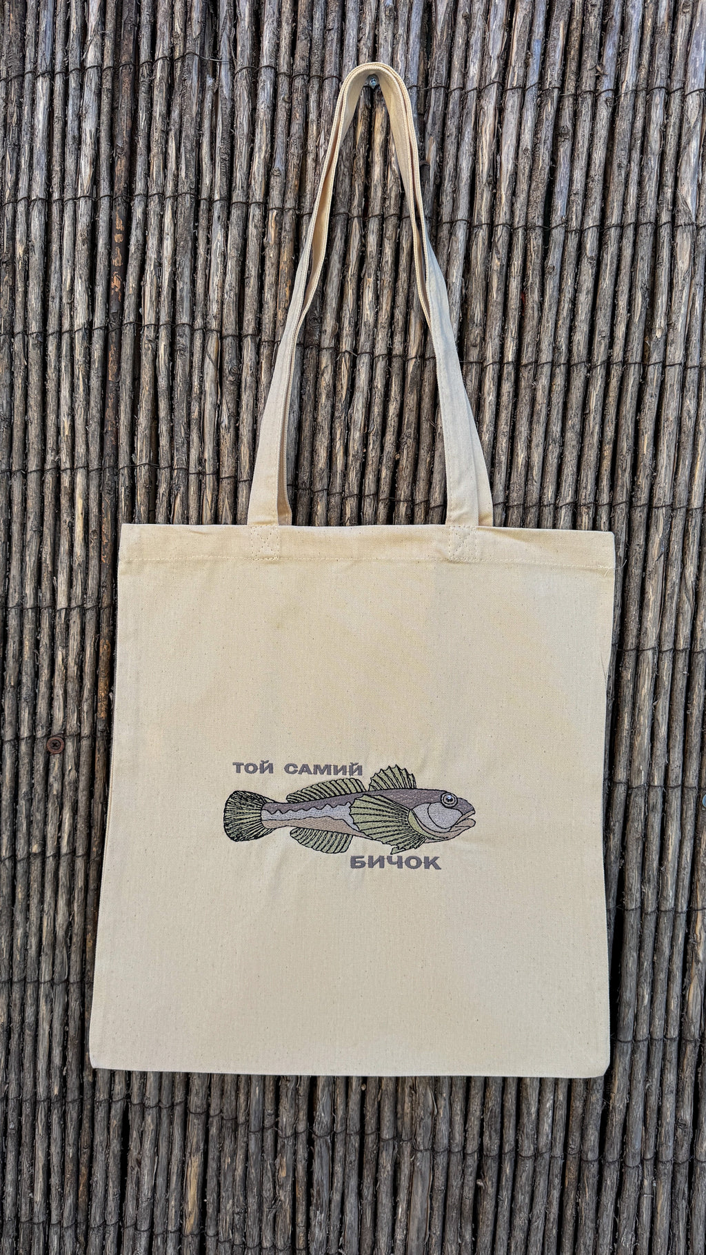 A natural beige canvas tote bag with long handles, featuring a printed illustration of a fish and Cyrillic text on the front, displayed against a dark brown woven bamboo background.