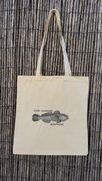 A natural beige canvas tote bag with long handles, featuring a printed illustration of a fish and Cyrillic text on the front, displayed against a dark brown woven bamboo background.