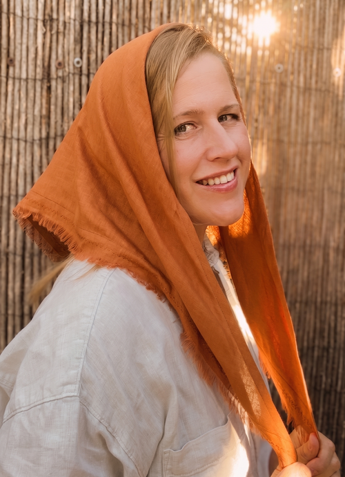 A smiling woman with blonde hair wearing a rust-orange headscarf and a light-colored linen shirt, posing in front of a warm, sunlit bamboo wall.