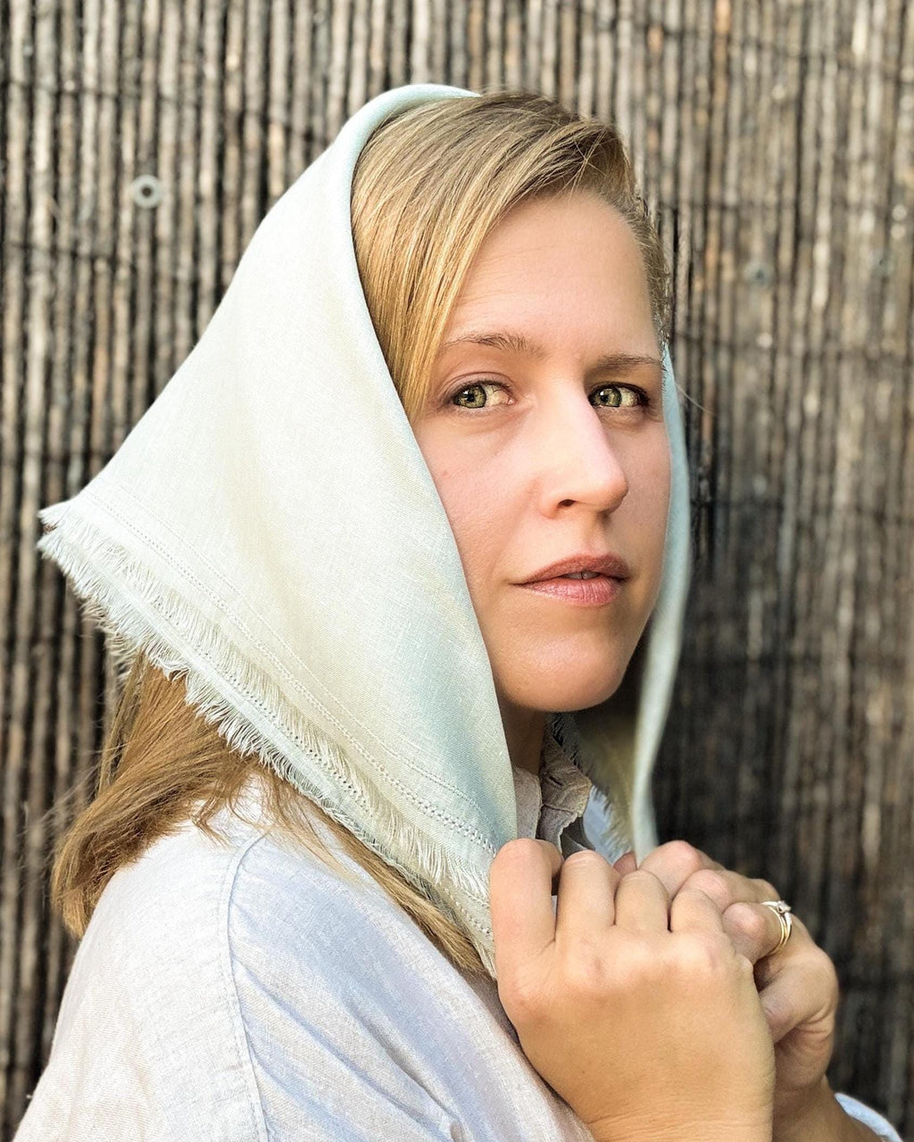 A woman wearing a light sage green fringed headscarf and a white shirt, posing in front of a rustic bamboo wall.