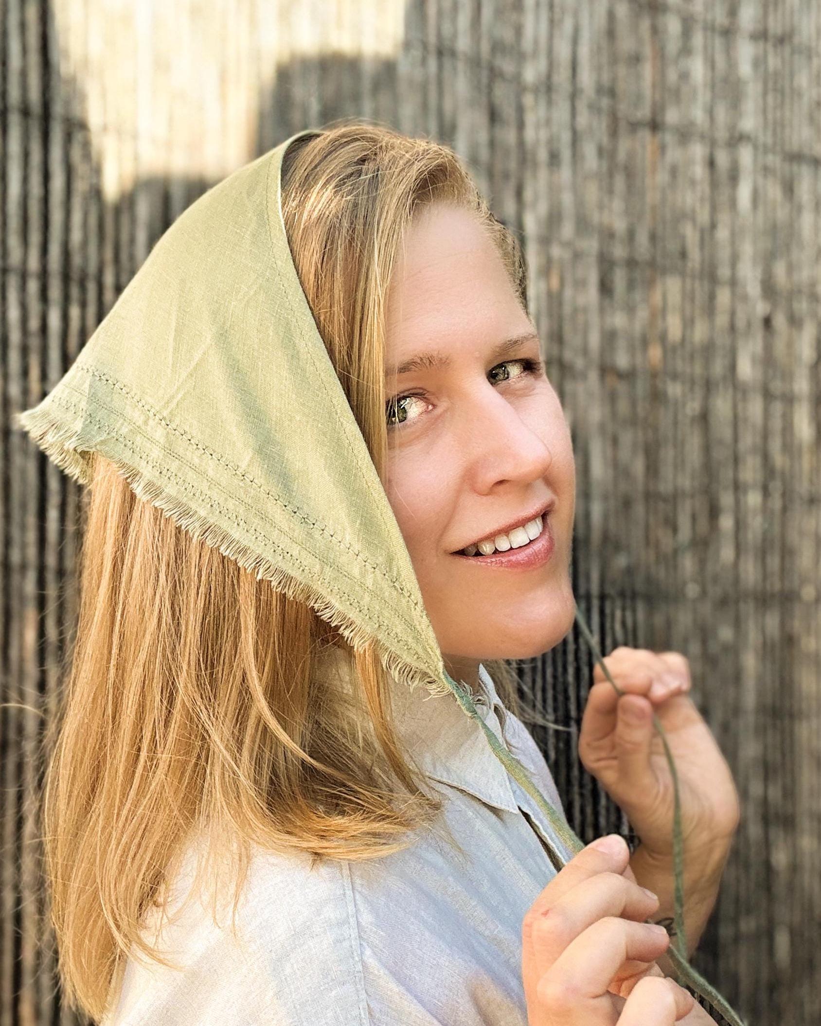 A smiling young woman wearing a light olive green fabric bonnet with frayed edges, holding a green string against a rustic wooden background.