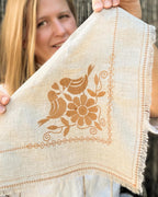 A woman holds up a square beige linen napkin with a brown embroidered design of two birds and a flower, featuring fringed edges and decorative borders.