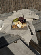 A rustic charcuterie board with cheese, grapes, and nuts sits on a weathered wooden table draped with a light beige linen cloth. A stack of white bowls is partially visible on the left, and a knife rests on the right, set against a background of a reed fence.