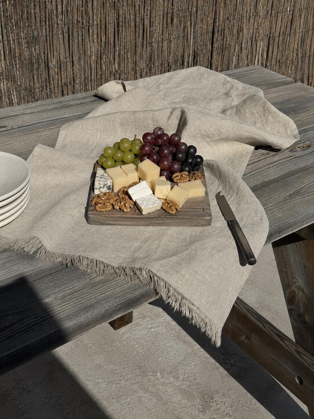 A rustic charcuterie board with cheese, grapes, and nuts sits on a weathered wooden table draped with a light beige linen cloth. A stack of white bowls is partially visible on the left, and a knife rests on the right, set against a background of a reed fence.