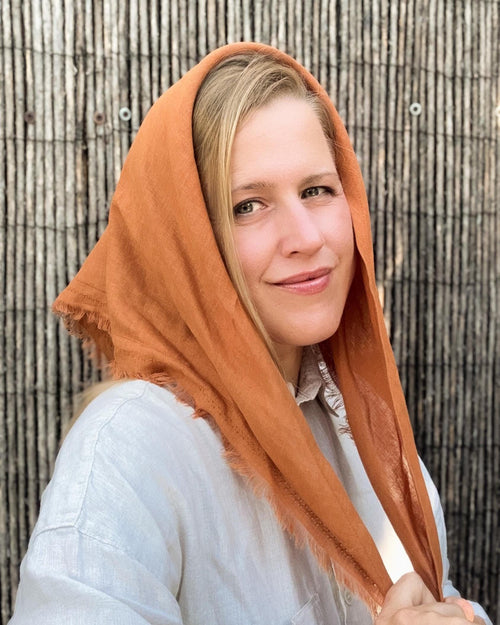 A woman wearing a rust-orange headscarf and a light-colored linen shirt, posing in front of a rustic wooden slat wall.