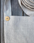 Close-up of a light gray linen tote bag with a coconut shell button closure and a matching woven strap, laid on a weathered gray wooden surface.