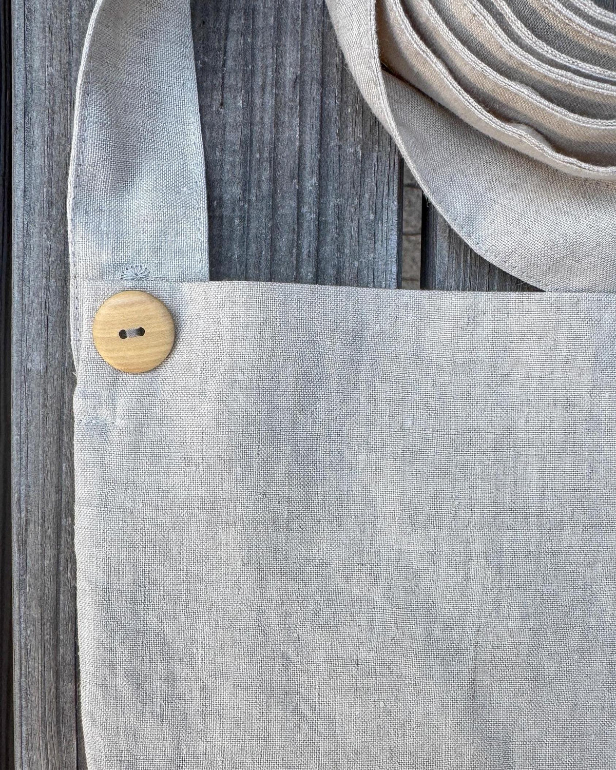Close-up of a light gray linen tote bag with a coconut shell button closure and a matching woven strap, laid on a weathered gray wooden surface.