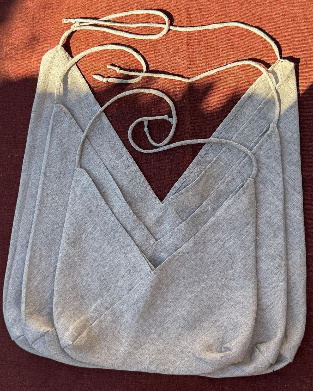 A light gray linen wrap-style shoulder bag with thin cream drawstrings, laid flat on a rust-colored fabric background.