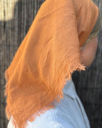 A close-up of a person wearing a draped terracotta orange linen scarf or shawl with frayed edges over the head and shoulders, paired with a light blue top, against a dark textured background.