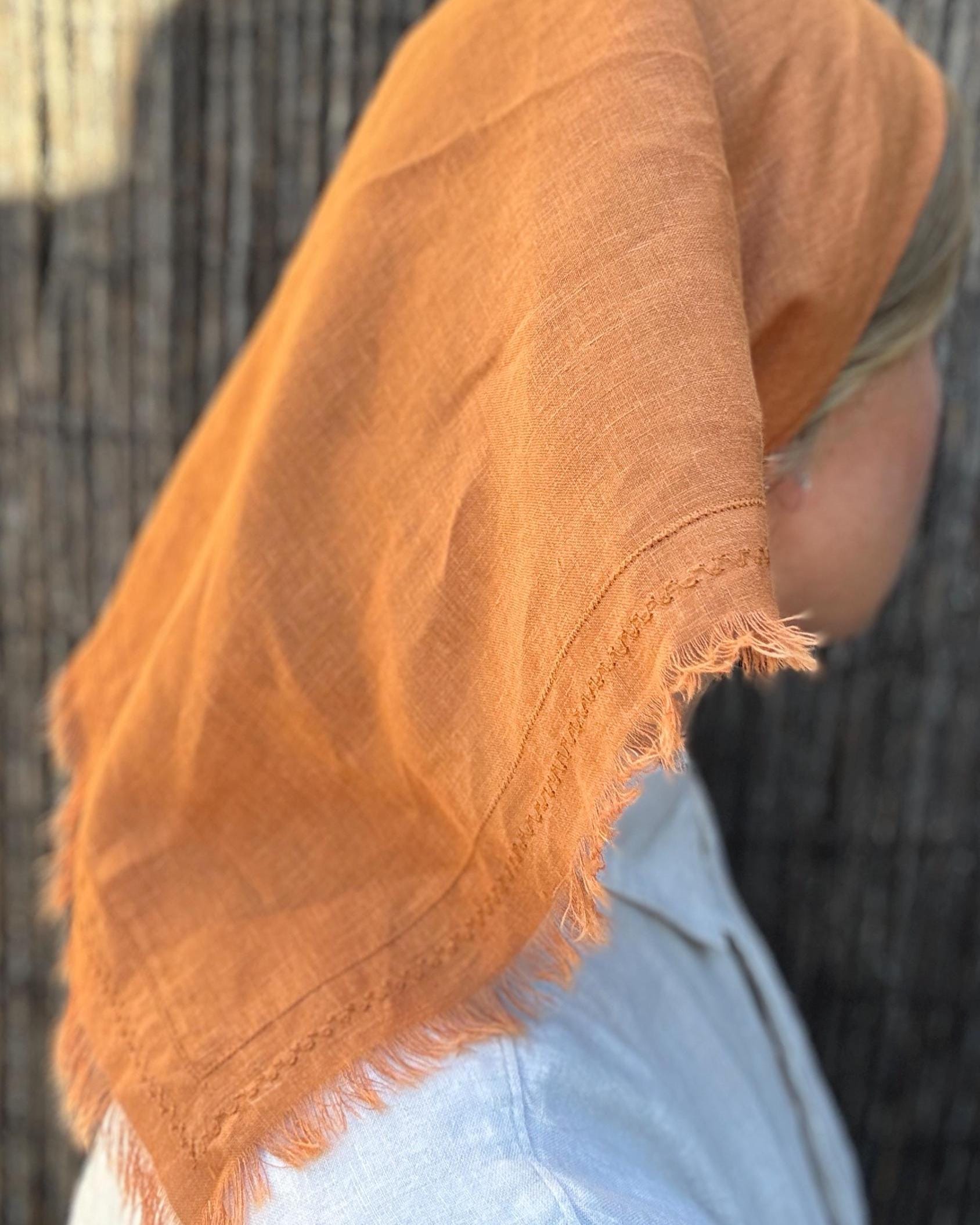A close-up of a person wearing a draped terracotta orange linen scarf or shawl with frayed edges over the head and shoulders, paired with a light blue top, against a dark textured background.