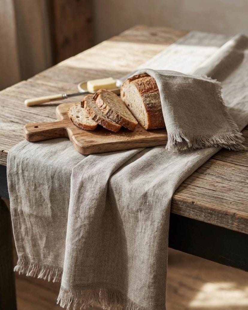 A light beige linen tablecloth with fringed edges draped over a rustic wooden table, styled with a wooden cutting board, sliced artisan bread, and a butter knife.