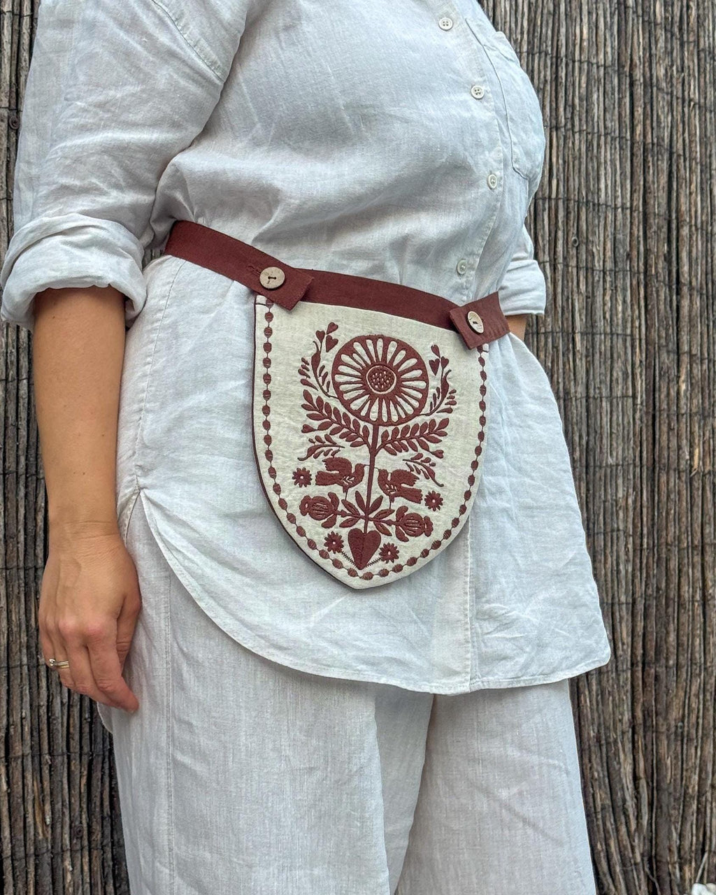 A person wearing a white linen shirt and matching trousers, with a brown linen waist strap and a cream shield-shaped pouch featuring a brown floral and sunburst print, standing against a bamboo wall.
