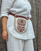 A person wearing a white linen shirt and matching trousers, with a brown linen waist strap and a cream shield-shaped pouch featuring a brown floral and sunburst print, standing against a bamboo wall.