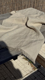 A light beige linen throw blanket with fringed edges draped over a dark wooden table outdoors on a paved surface.