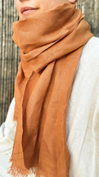 A close-up of a person wearing a draped terracotta orange scarf wrapped around their neck and shoulders, paired with a light-colored linen top against a bamboo background.