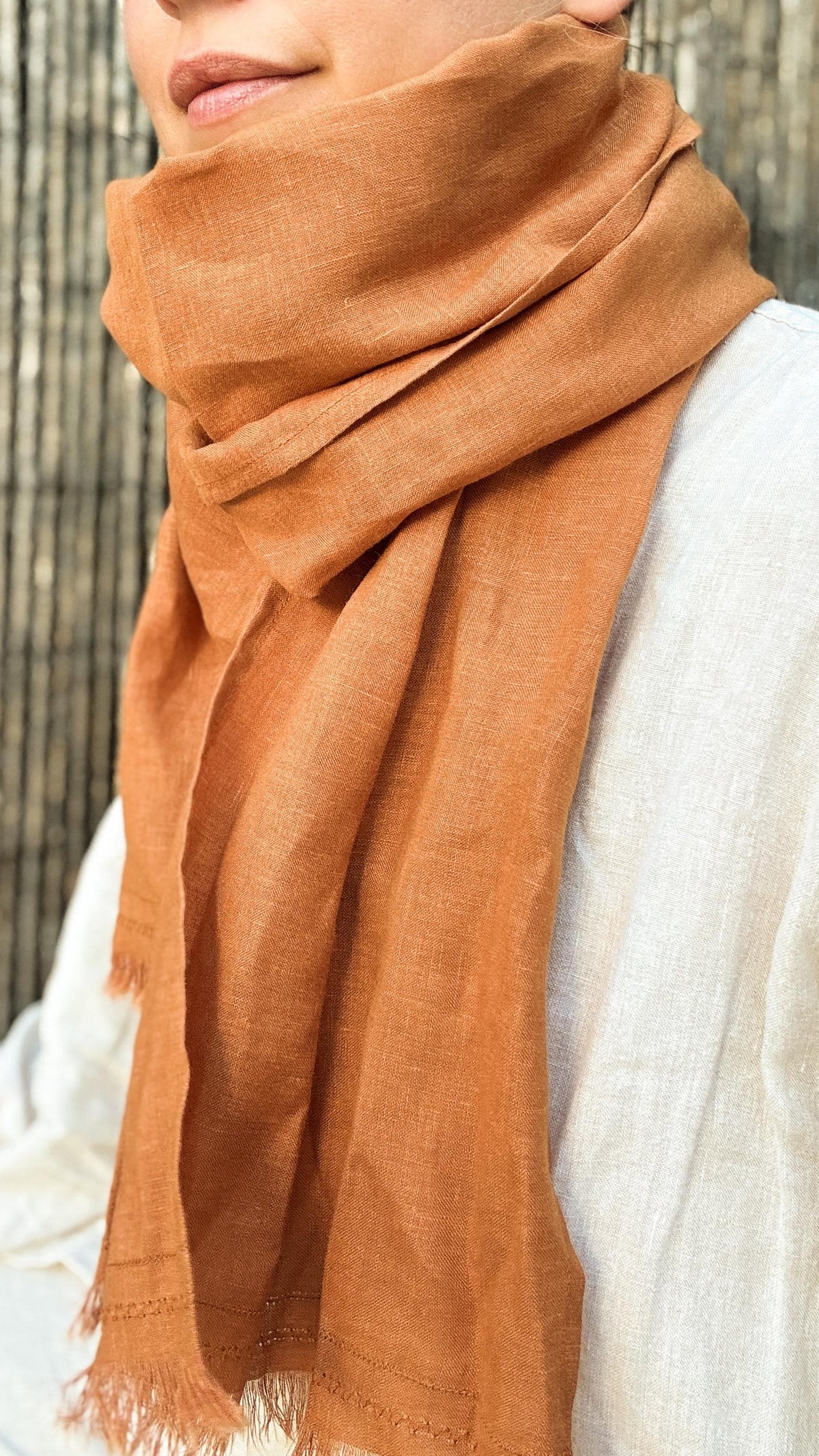A close-up of a person wearing a draped terracotta orange scarf wrapped around their neck and shoulders, paired with a light-colored linen top against a bamboo background.