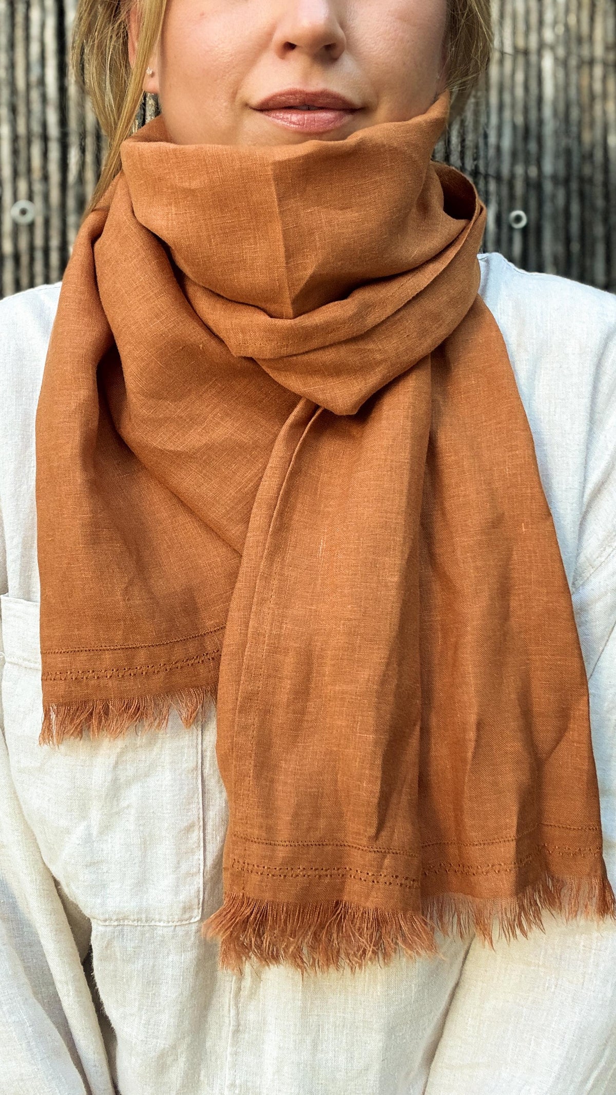 A close-up of a person wearing a warm rust-orange scarf wrapped around their neck over a light-colored linen top, with a rustic wooden fence in the background.