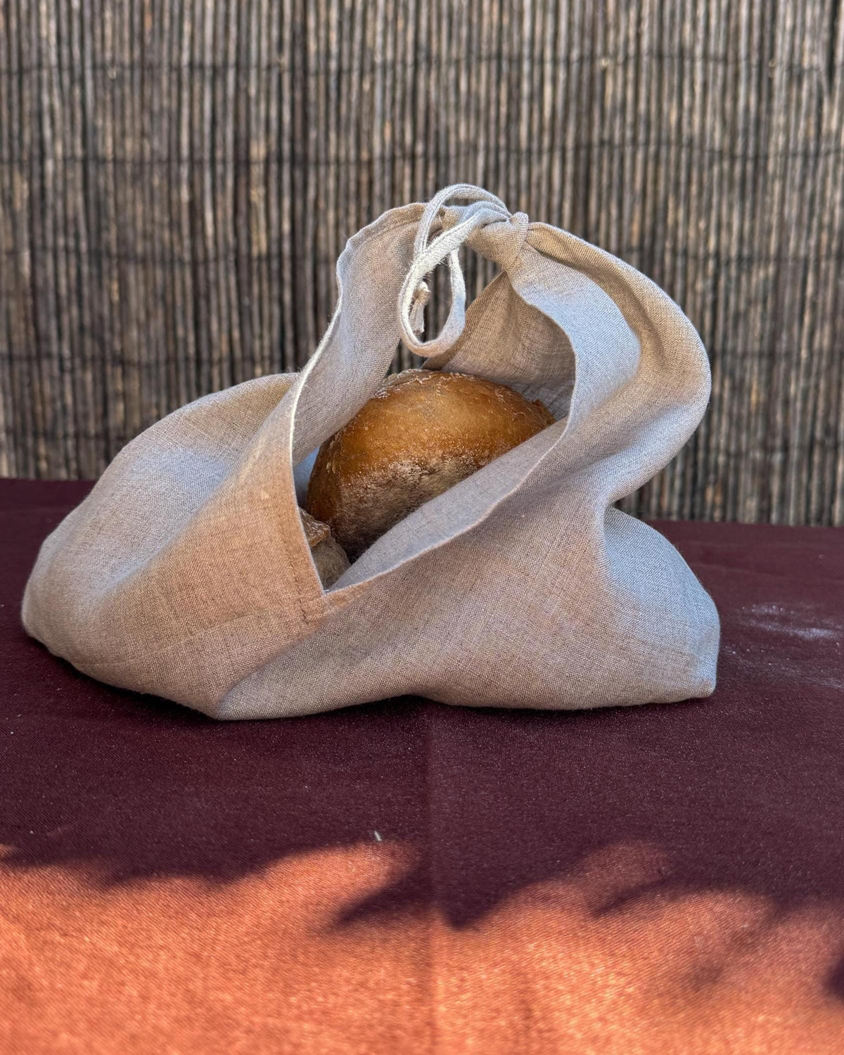 A beige linen drawstring bag holding a round brown item, placed on a dark red surface with a woven brown background.