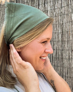 A woman wearing an olive green fabric head covering or bandana-style hair wrap, shown in profile against a rustic bamboo or reed wall.