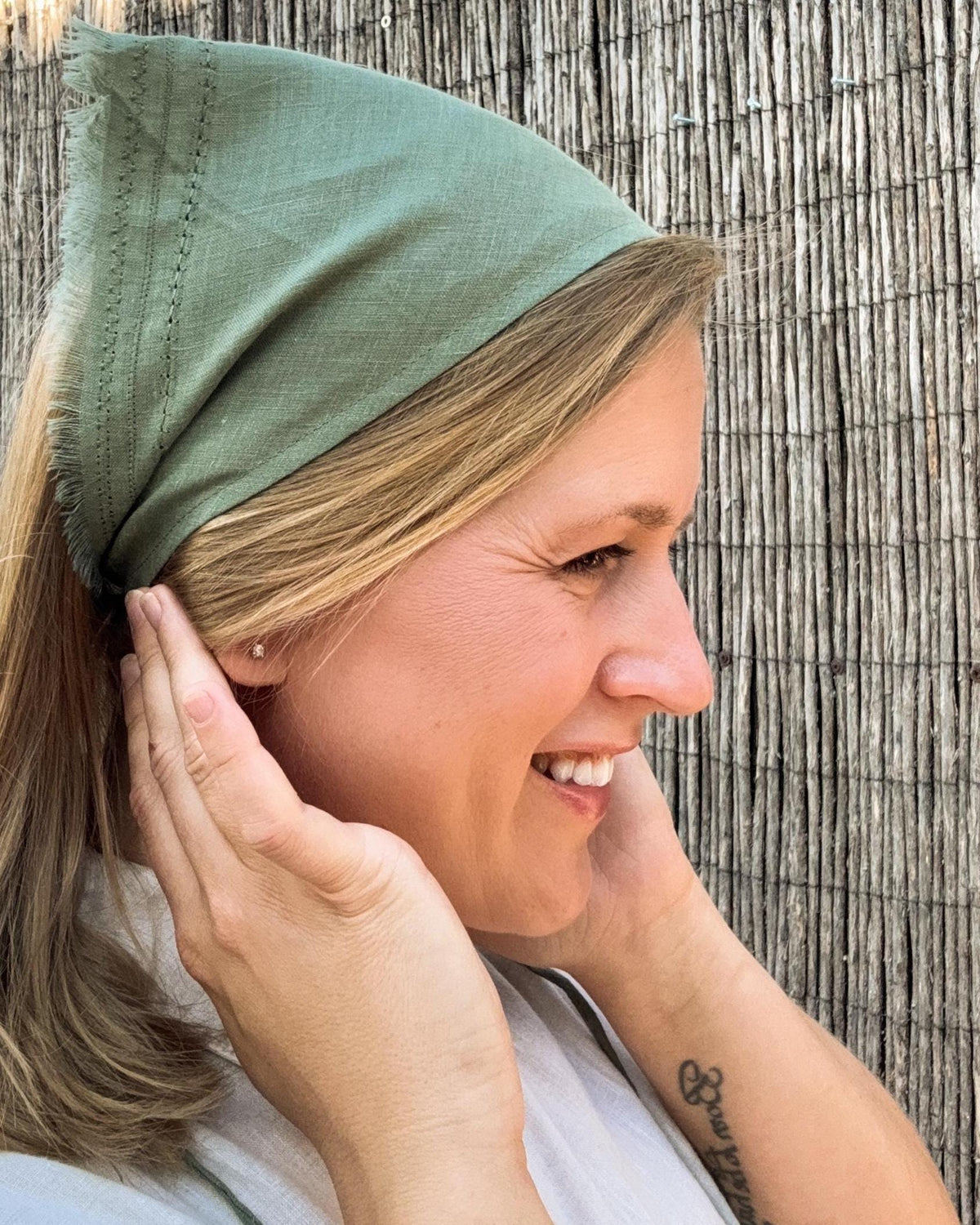 A woman wearing an olive green fabric head covering or bandana-style hair wrap, shown in profile against a rustic bamboo or reed wall.