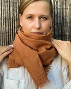 A woman wearing a warm rust-orange textured scarf with fringed edges, styled around her neck over a white linen shirt, set against a dark bamboo background.