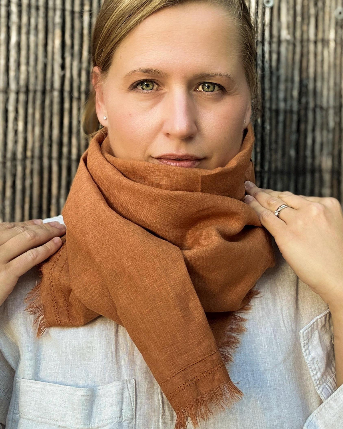 A woman wearing a warm rust-orange textured scarf with fringed edges, styled around her neck over a white linen shirt, set against a dark bamboo background.