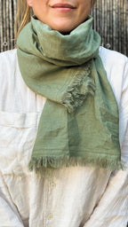 A sage green linen scarf with fringed edges, wrapped around the neck of a person wearing a white linen shirt.