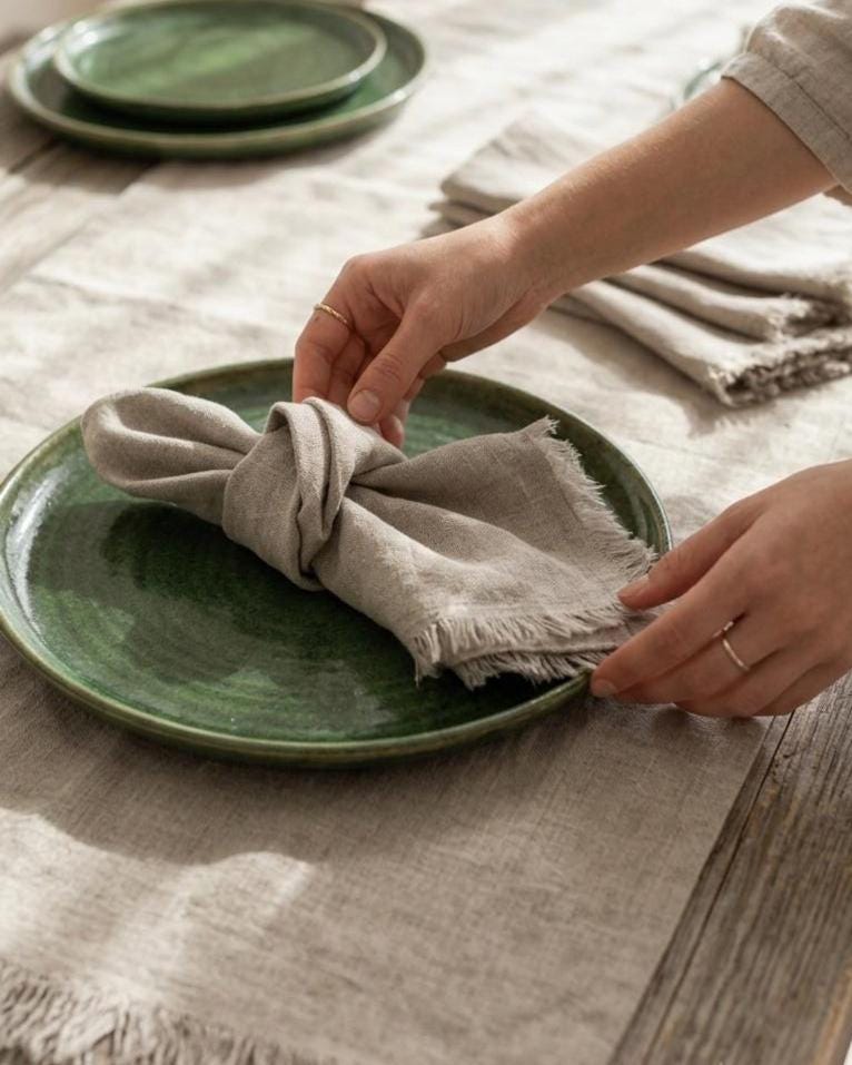 A close-up of a person's hands arranging a light beige linen napkin on a round green ceramic plate, set on a textured beige tablecloth with matching green plates in the background.