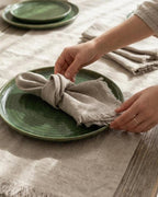 A close-up of a person's hands arranging a light beige linen napkin on a round green ceramic plate, set on a textured beige tablecloth with matching green plates in the background.