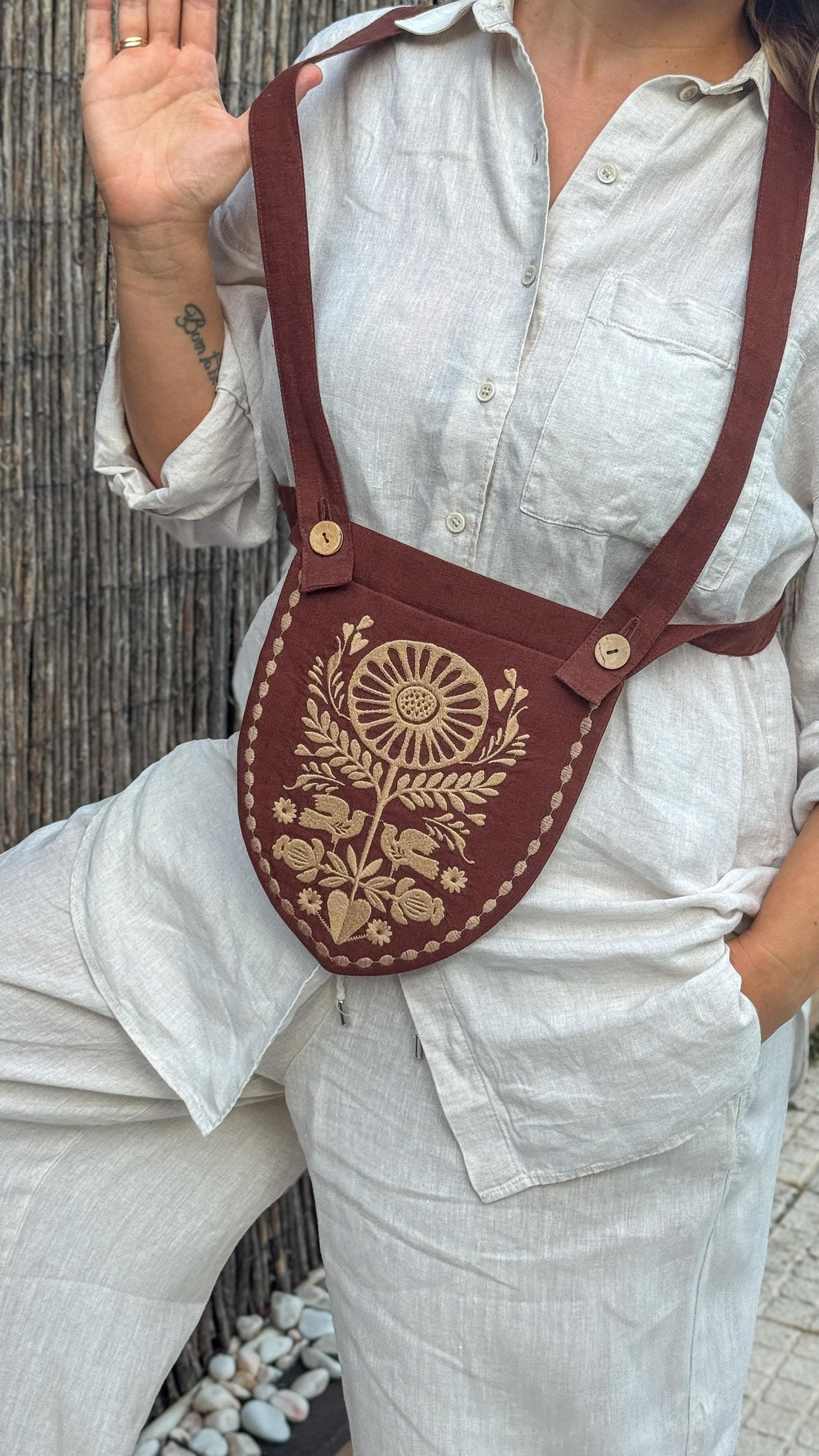 A person wearing a white linen shirt and matching pants carries a brown linen shoulder bag with a large beige floral sunburst design on the front.