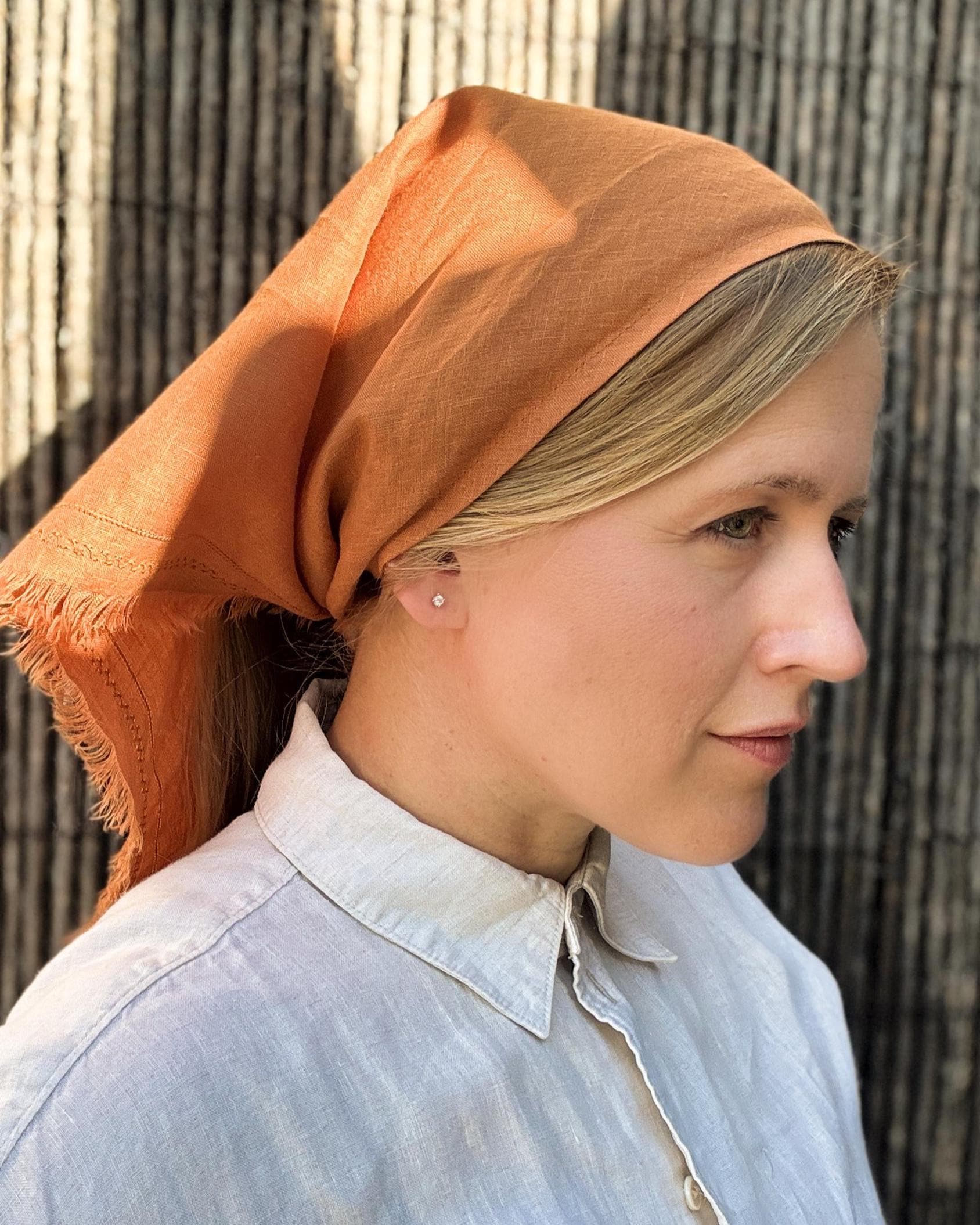 A woman in profile wearing a terracotta orange linen headscarf and a light blue linen button-up shirt, set against a rustic bamboo screen background.
