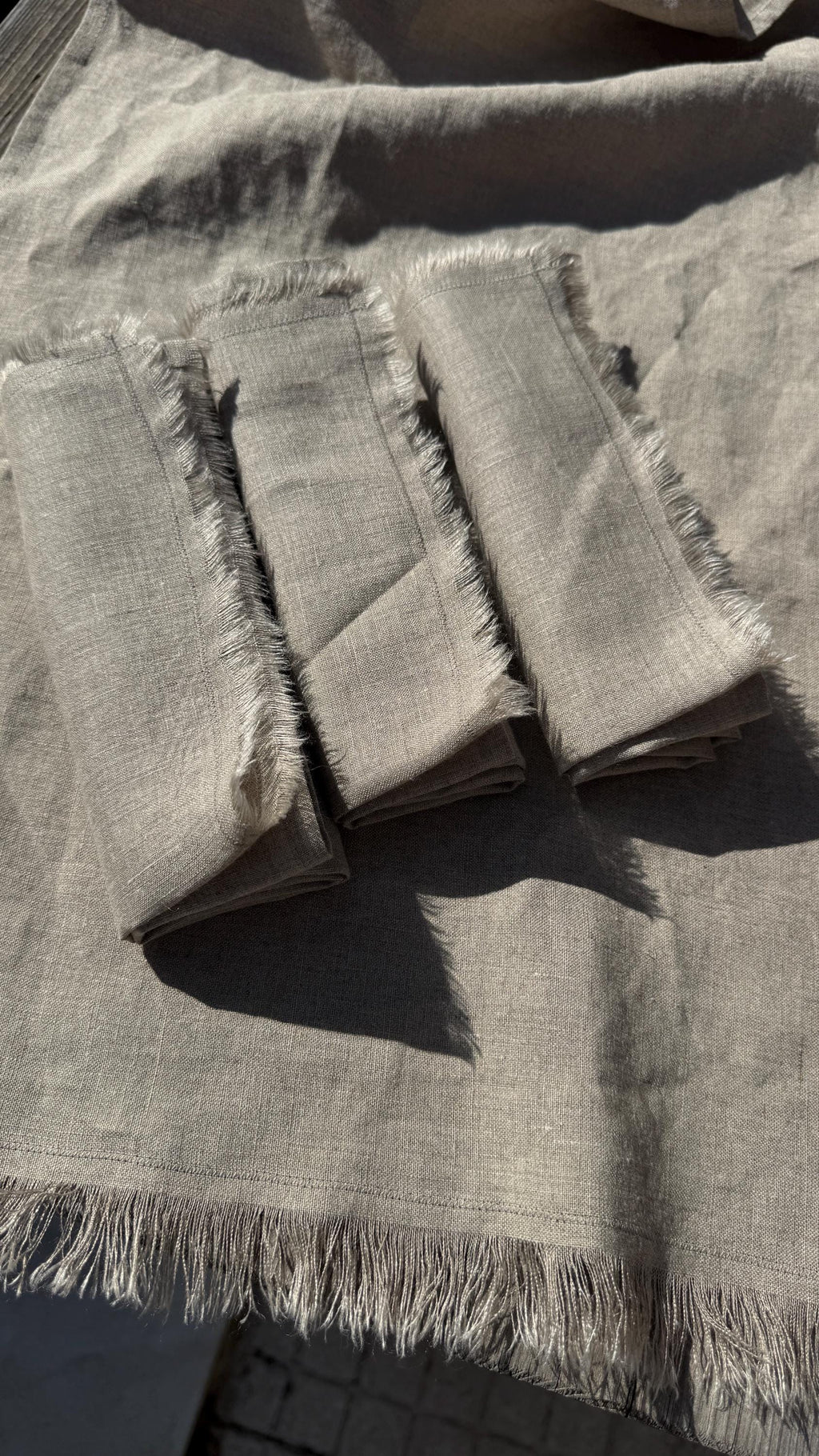 Three folded square fabric napkins with frayed edges laid on top of a matching light gray linen cloth, photographed in bright natural sunlight.