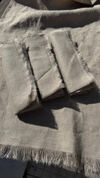 Three folded square fabric napkins with frayed edges laid on top of a matching light gray linen cloth, photographed in bright natural sunlight.