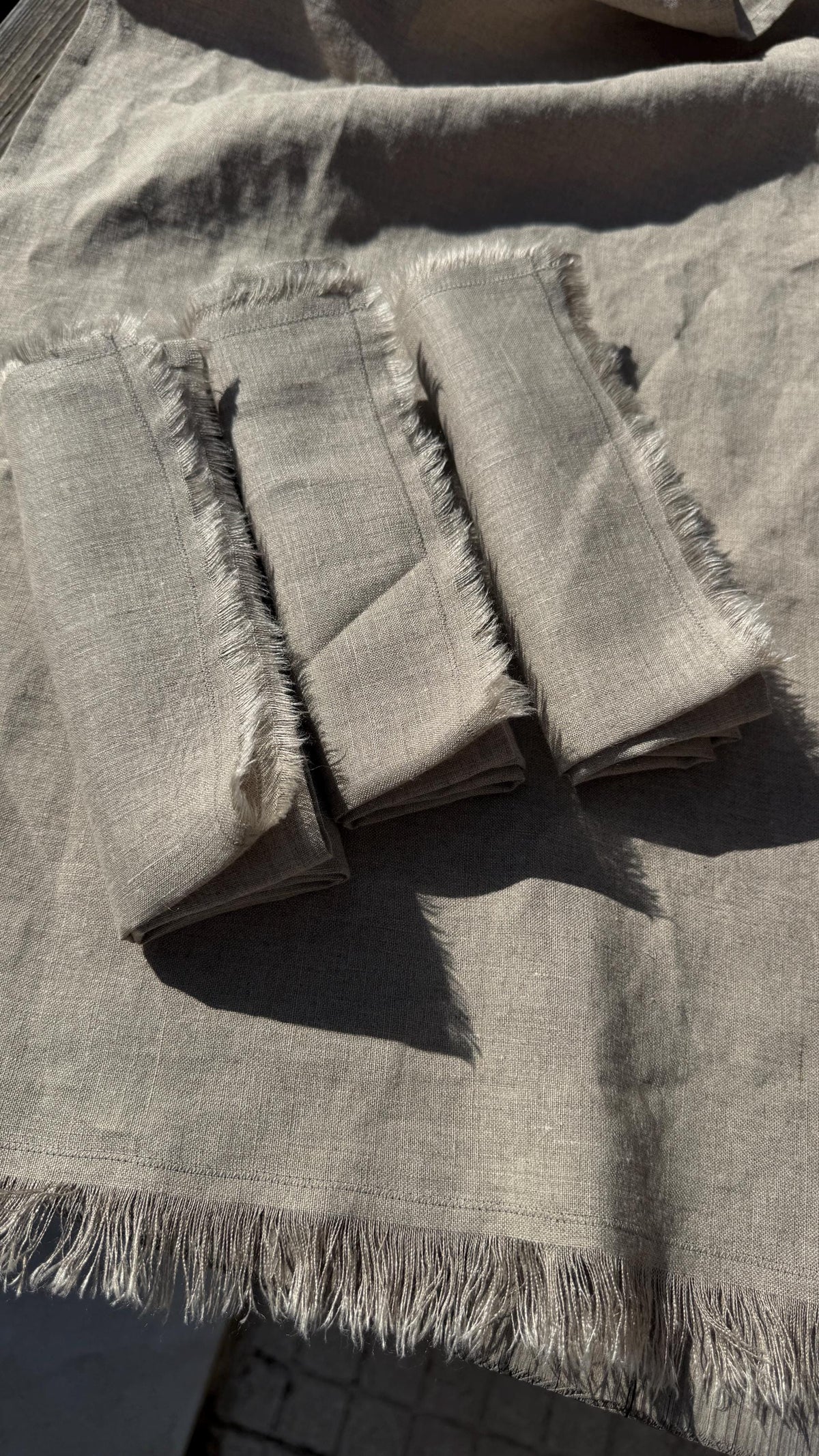 Three folded square fabric napkins with frayed edges laid on top of a matching light gray linen cloth, photographed in bright natural sunlight.